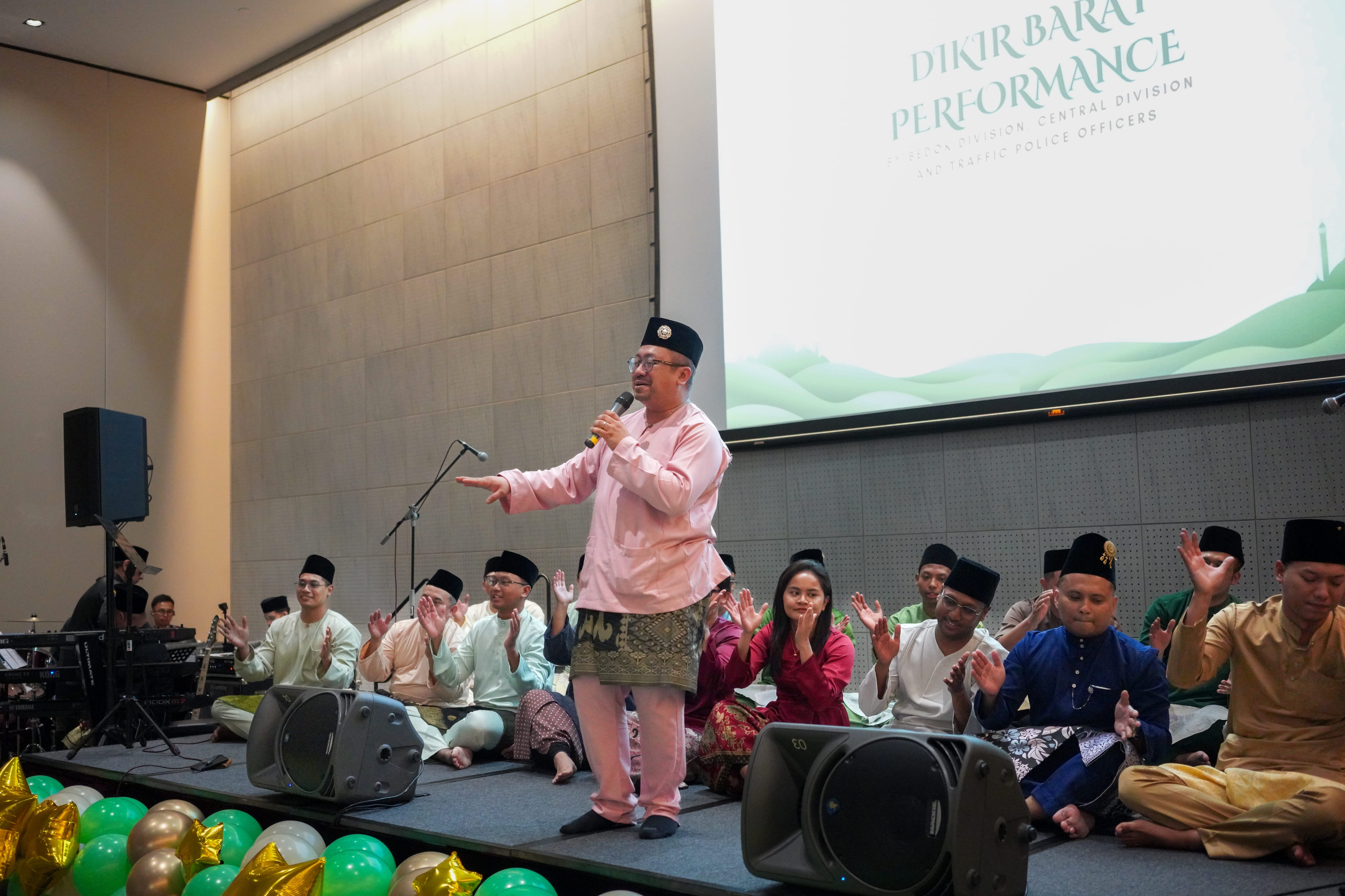  performer speaking into a microphone on stage during a dikir barat performance, with fellow performers seated behind him.